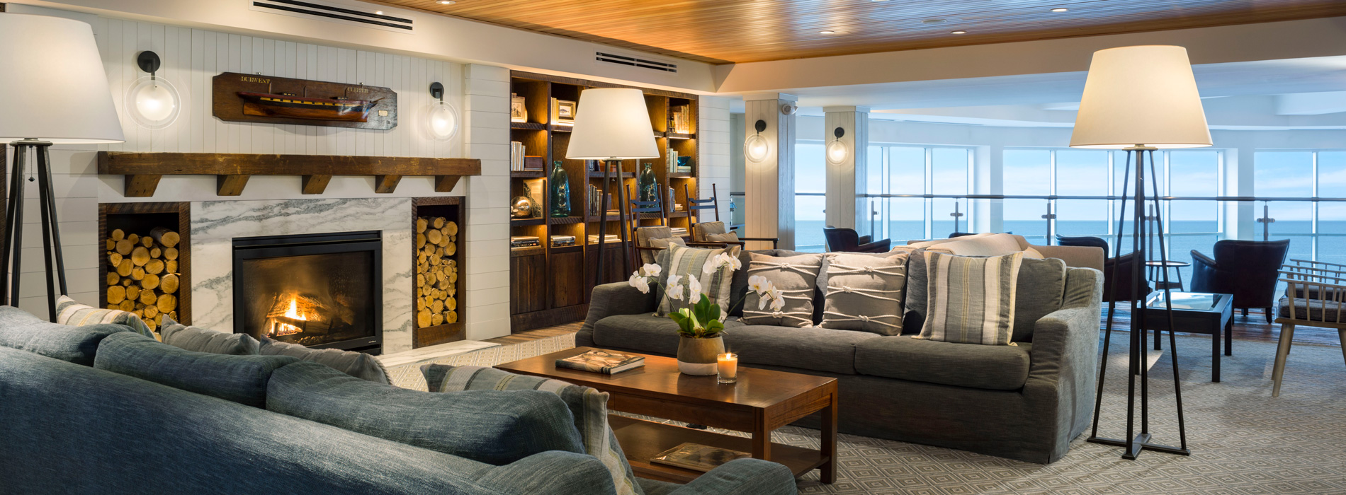 The lobby at Cliff House Resort in Maine with cozy couches surrounding a wood burning fireplace and walls of books with the ocean revealed in the wall of windows beyond.